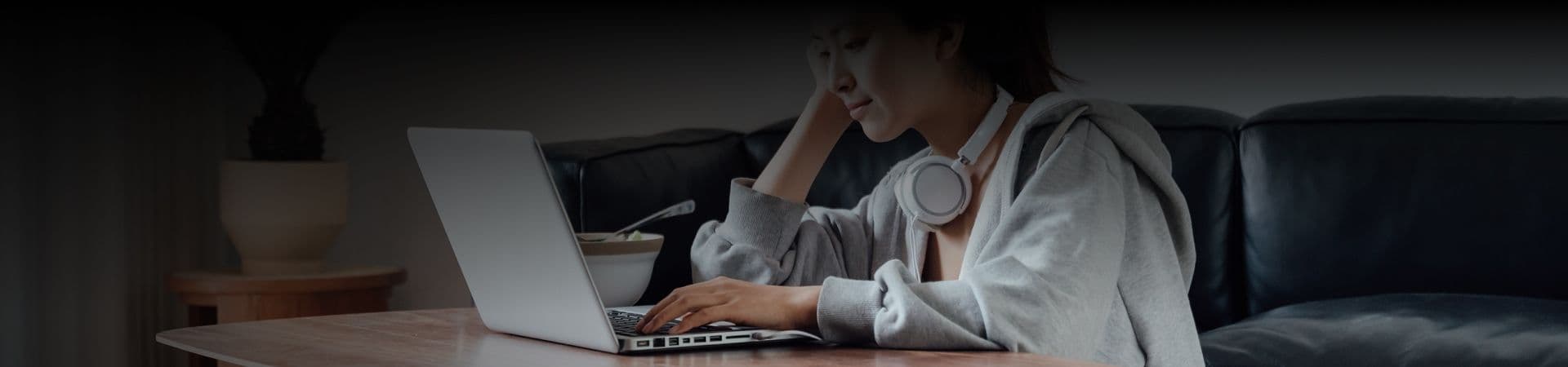 An image of a woman working on her laptop wearing a grey hoodie and headphones around her neck.