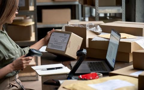 An image shows a woman checking information on a courier box while holding a barcode scanner in her hand.