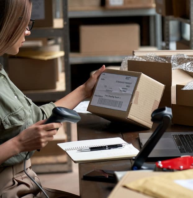 An image shows a woman checking information on a courier box while holding a barcode scanner in her hand.